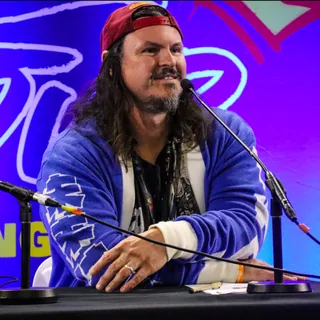 Mike Roberts, smiling at a panel desk in a backwards cap and blue cardigan