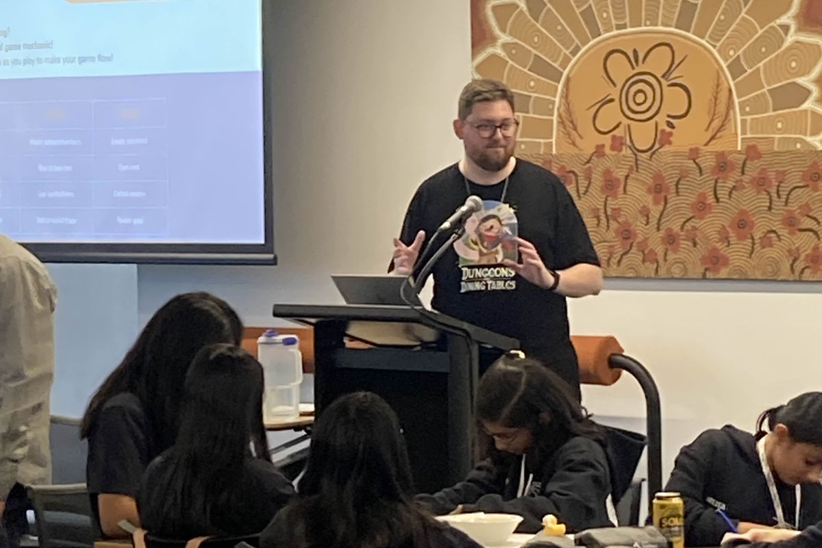 A Catalyst Games facilitator presents game-design mechanics to a room of secondary-school students working at tables during a workshop.